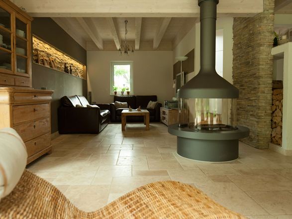 Woonkamer met vrijstaande open haard en zithoek op Lichte travertin tegels, een rieten fauteuil op de voorgrond